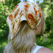 A blonde woman shown from the back outdoors, wearing a cream-colored silk-like headscarf with a rust-orange floral pattern.