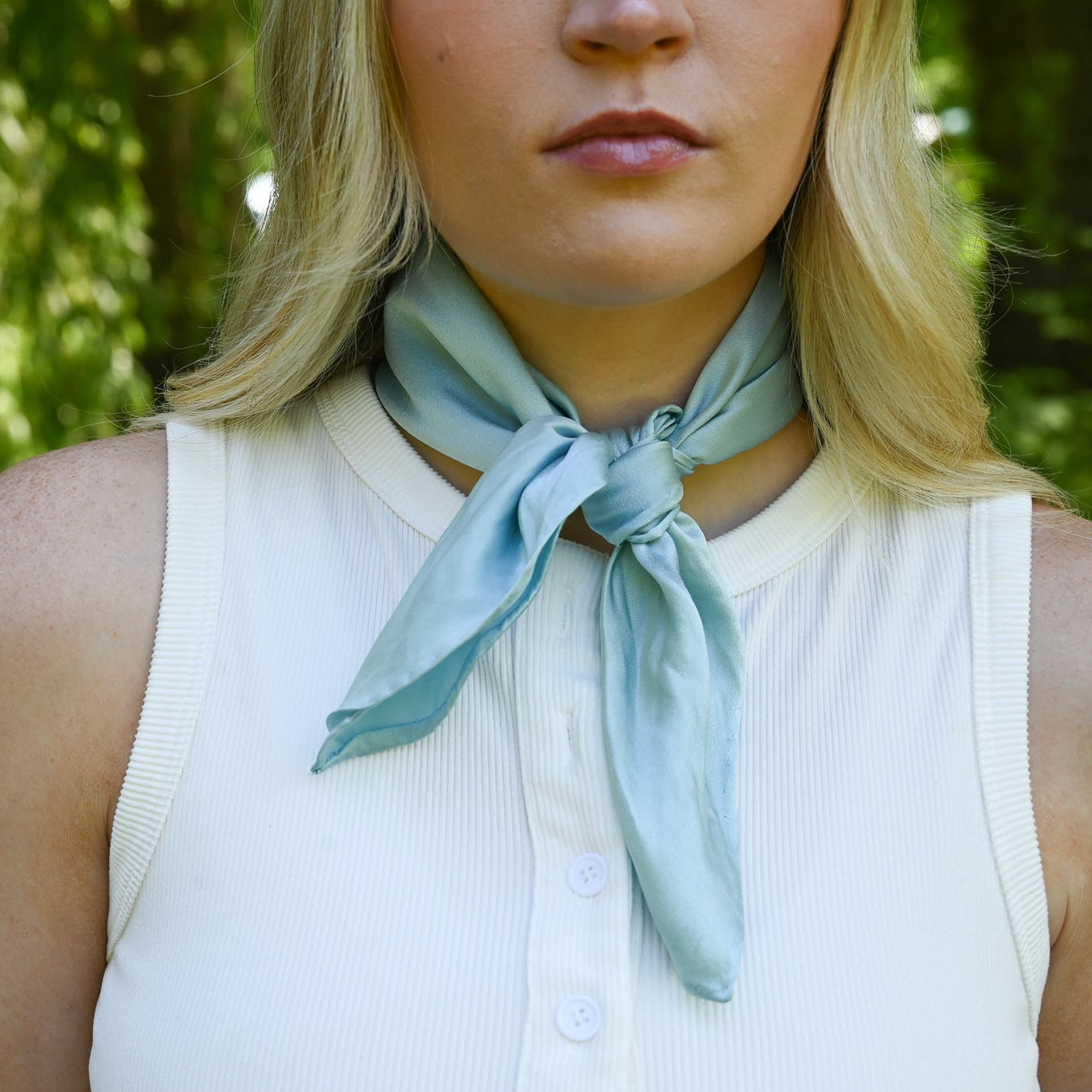 A close-up of a woman outdoors wearing a light blue satin neck scarf tied in a knot over a white sleeveless top, with a blurred green forest background.