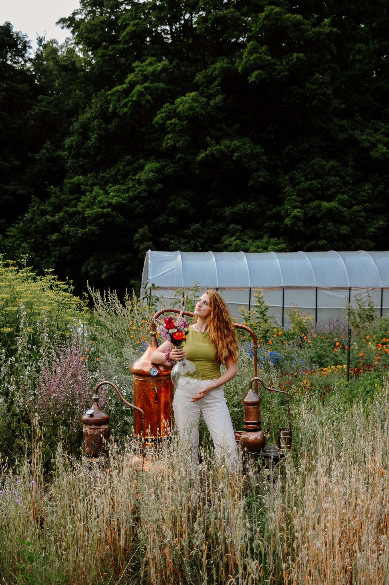 Woman standing in a garden next to three copper stills with a greenhouse in the background