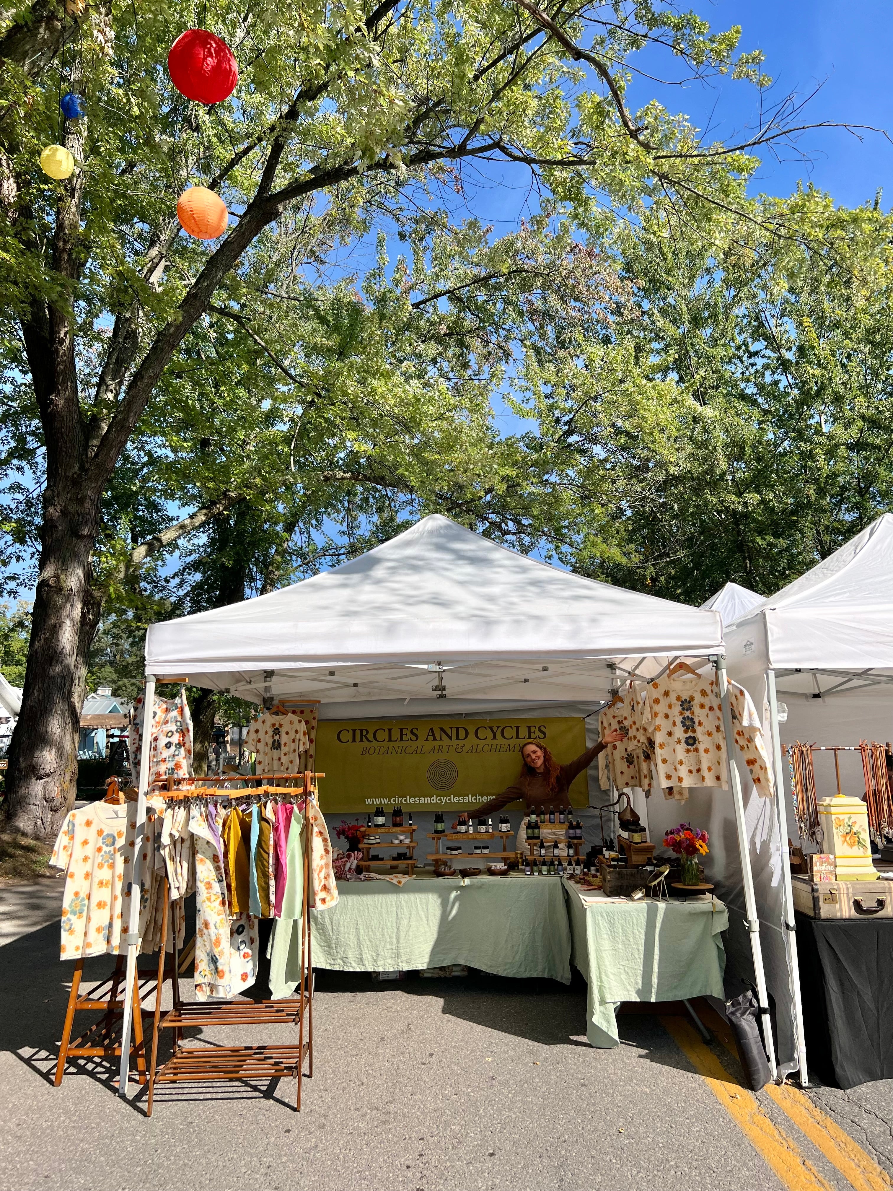 Outdoor market stall with a white tent, tables, and various items on display under a tree.