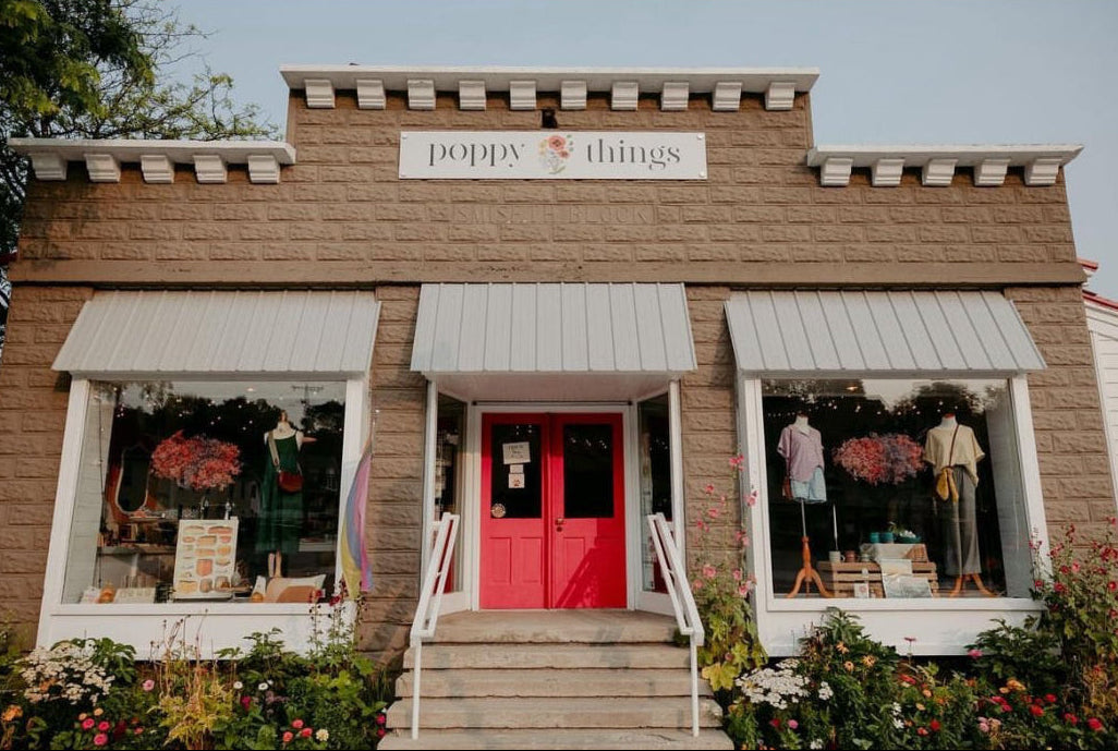 Storefront with a red door and 'poppy things' sign, surrounded by greenery.