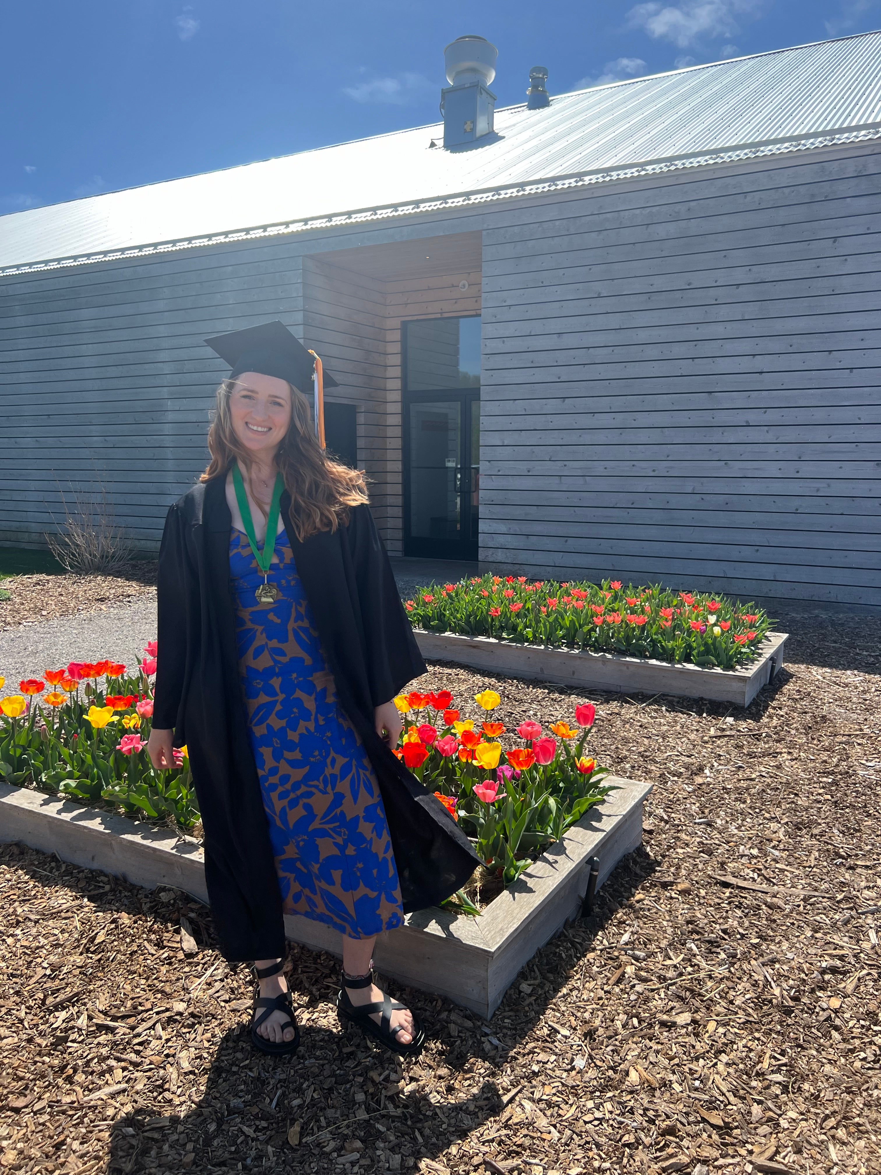 Graduate in cap and gown standing in front of a building with tulip flowers.