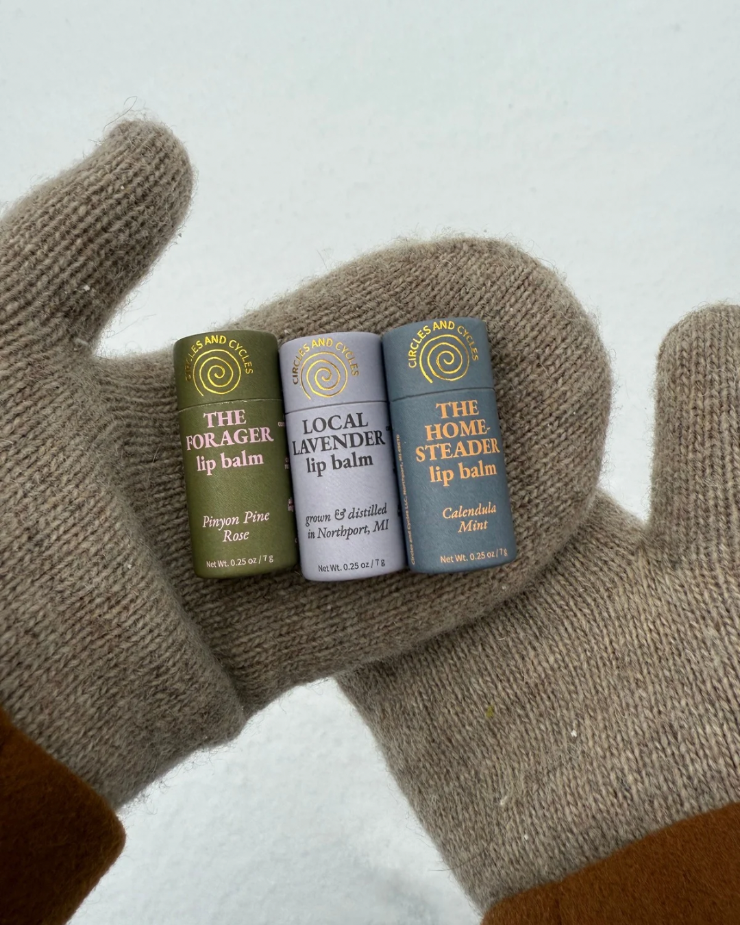 Three lip balm tubes held in a gloved hand against a light background